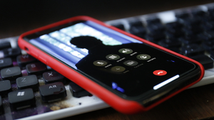 Na zdjęciu widoczny jest telefon z etui/obudowa koloru czerwonego leżący na klawiaturze komputera. Na ekranie smartfon widać sylwetkę osoby podczas połączenia wideo.