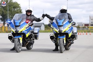 Zdjęcie przedstawia dwóch policjantów pionu ruchu drogowego ubranych w kombinezony policyjne siedzących na motocyklach służbowych oznakowanych policyjnych koloru srebrnego z niebieskimi elementami. Motocykliści  stykają się dłońmi