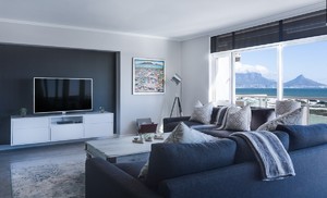 Grafika przedstawia wizualizację salonu/apartamentu z kanapą koloru szarego w kształcie litery L, telewizorem i szafką RTV koloru białego i dużym oknem z widokiem na góry. Na szarej podłodze jest rozłożony jasny prostokątny dywan na którym stoi stolik kawowy.