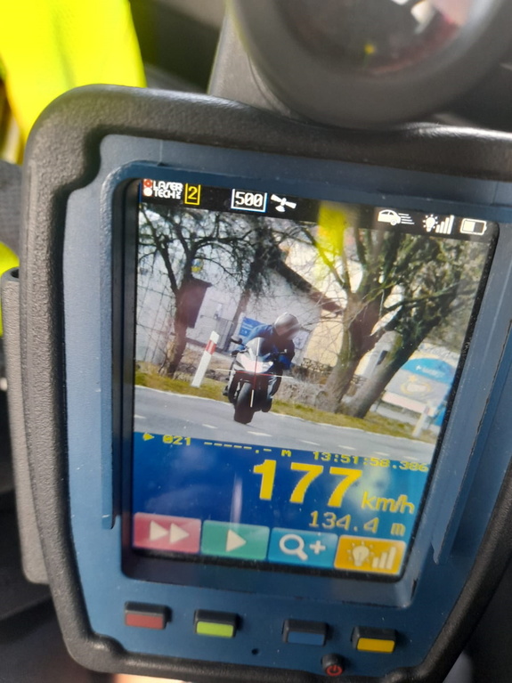 Zdjęcie przedstawia ekran miernika prędkości na którym widoczny jest jadący drogą kierujący motocyklem koloru niebieskiego
